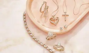 A collection of diamond jewelry displayed on a pink tray. The jewelry includes a diamond tennis bracelet, diamond hoop earrings, a heart-shaped diamond necklace, a diamond cross necklace, diamond stud earrings, and two diamond engagement rings. The image highlights the beauty and elegance of diamond jewelry.