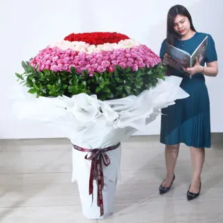 A grand bouquet of red, pink, and peach roses with green leaves in a white wrap