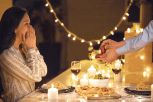 10 Gift Ideas to Kick Off the Valentine's Week in Style 6 At a dinner table, a man offers a ring to a woman, symbolizing a heartfelt proposal in an intimate setting
