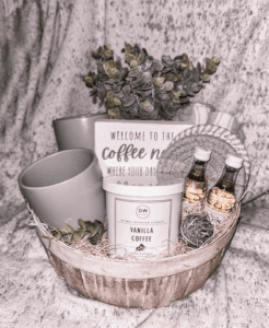 A cozy coffee lover's gift basket filled with a variety of items: a gray mug, a vanilla coffee candle, a welcome sign, and small bottles of hand sanitizer. The basket is decorated with greenery and nestled on a soft gray blanket.