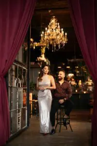 A couple stands happily in a restaurant, framed by the glow of an ornate chandelier above them.