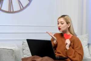 A woman sitting on a couch blows a kiss towards a laptop screen, holding a red paper heart in her other hand.