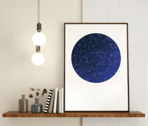 A framed poster depicting a circular map of the night sky, showcasing constellations and stars. The poster is hanging on a wall above a wooden shelf with books, a vase, and a hanging light.