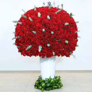 A large arrangement of 1500 red roses with eucalyptus in a white vase surrounded by greenery at the bottom.