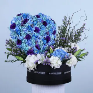 A Flower box featuring white and blue hydrangeas, roses, lisianthus, and aster arranged in a heart shape in a round black box.