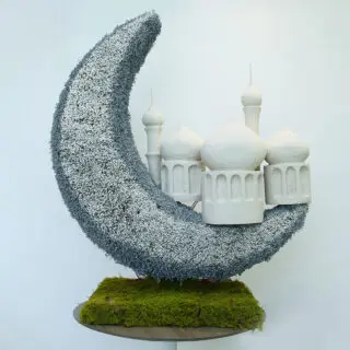 A grand crescent-shaped grey floral arrangement with a miniature white mosque, set on a moss-covered base on top of a sleek white stand.