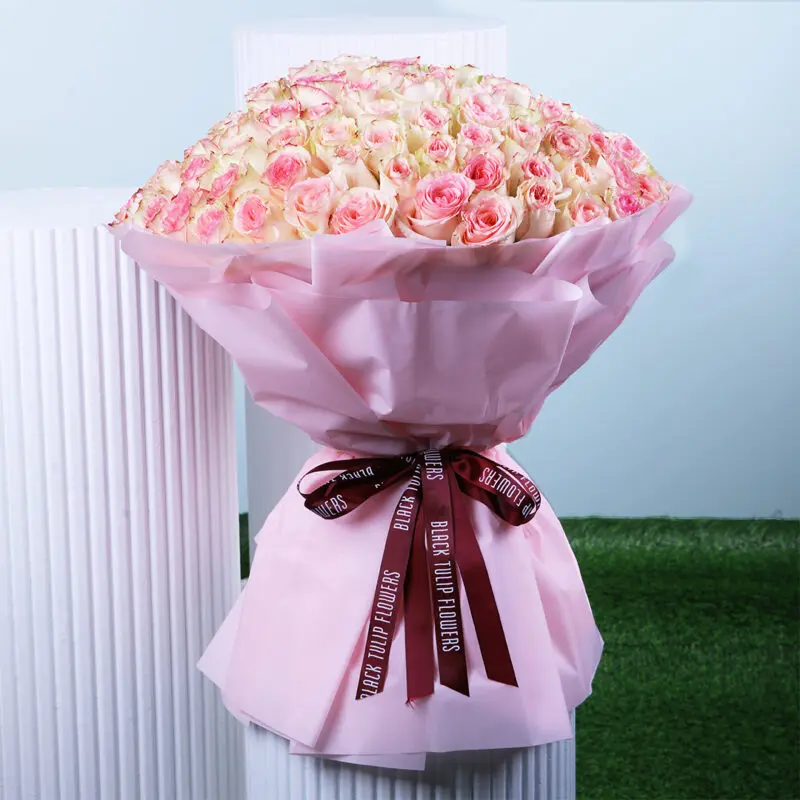 A beautifully arranged bouquet of fresh roses in a soft pink wrap, tied with a maroon ribbon.