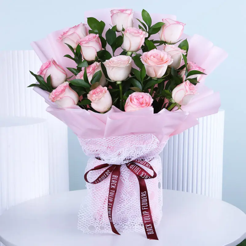 Blush Rose Bouquet A beautiful bouquet featuring soft pink roses, hand-tied and wrapped in light pink, displayed on a white table.