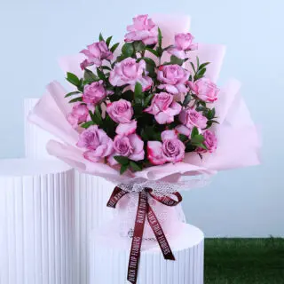 A bouquet of roses and greenery, hand-tied and wrapped in light pink tied with maroon ribbon displayed on a white pedestal.