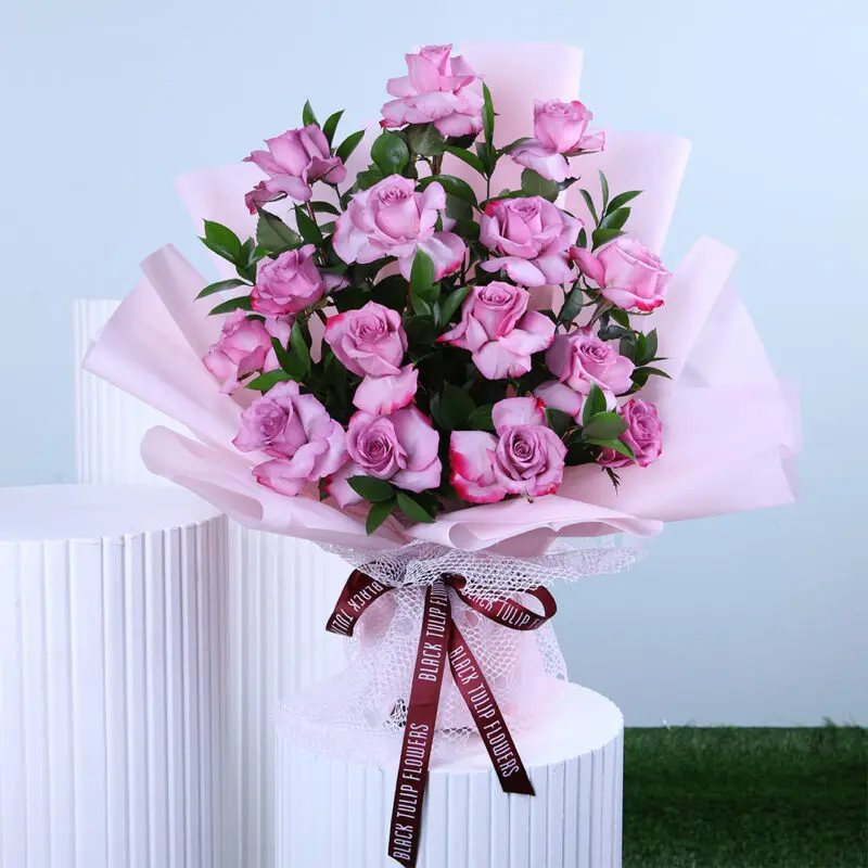 Graceful Rose Bouquet A bouquet of roses and greenery, hand-tied and wrapped in light pink tied with maroon ribbon displayed on a white pedestal.