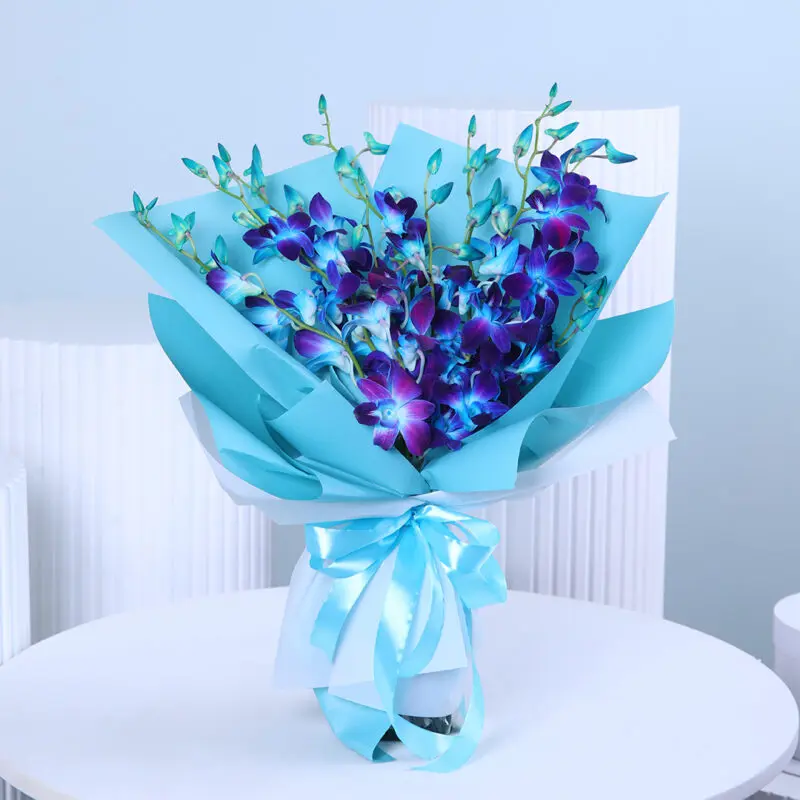 blue-orchid-bouquet A hand-tied bouquet of vibrant blue dendrobium orchids with fresh green leaves, wrapped in light teal paper and tied with a light blue ribbon.