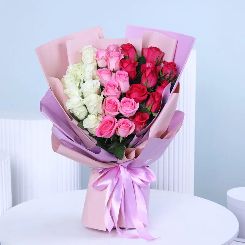 A bouquet of white, pink, and red roses clustered and wrapped in layers of light pink and lavender paper, tied with a wide, light purple satin ribbon.