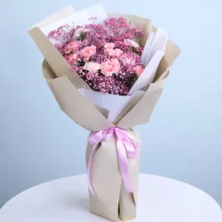 A bouquet of light pink carnations and pink baby's breath wrapped in brown kraft paper with layers of white tissue and tied with a light pink satin ribbon.