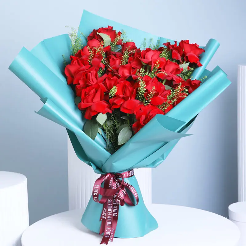 red-roses-bouquet A premium bouquet of deep red roses with green foliage and green wrap, tied with a ribbon, set against a soft blue background.
