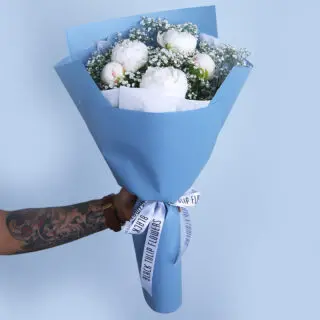 Elegant bouquet of white peonies and baby’s breath wrapped in soft blue paper and tied with a white ribbon.