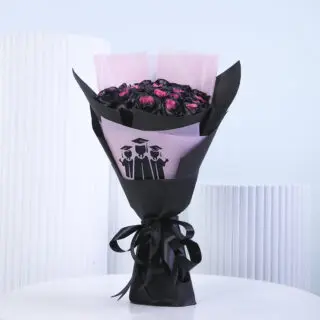 A bouquet of 30 roses with black-painted outer petals and visible pink centers, wrapped in black and pink paper. The pink wrapping has a black illustration of three graduation cap figures tied with a black ribbon.