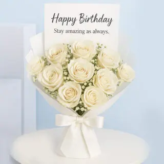 A bouquet of Cream roses with delicate baby's breath, wrapped in white paper and tied with a satin ribbon, with a "Happy Birthday, Stay amazing as always." card.