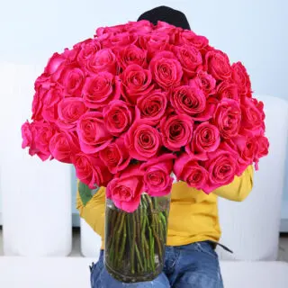 A large, full bouquet of 100 purple roses with visible green stems, meticulously arranged in a tall, clear glass cylindrical vase and tied with a natural-colored fabric.