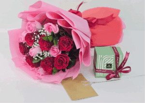 Pink and red rose bouquet wrapped in pink paper, placed beside a gift box and a golden envelope.