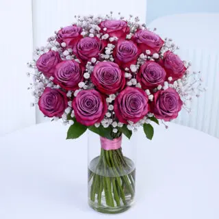 Deep purple rose floral arrangement with white baby’s breath in a glass vase on a white table.