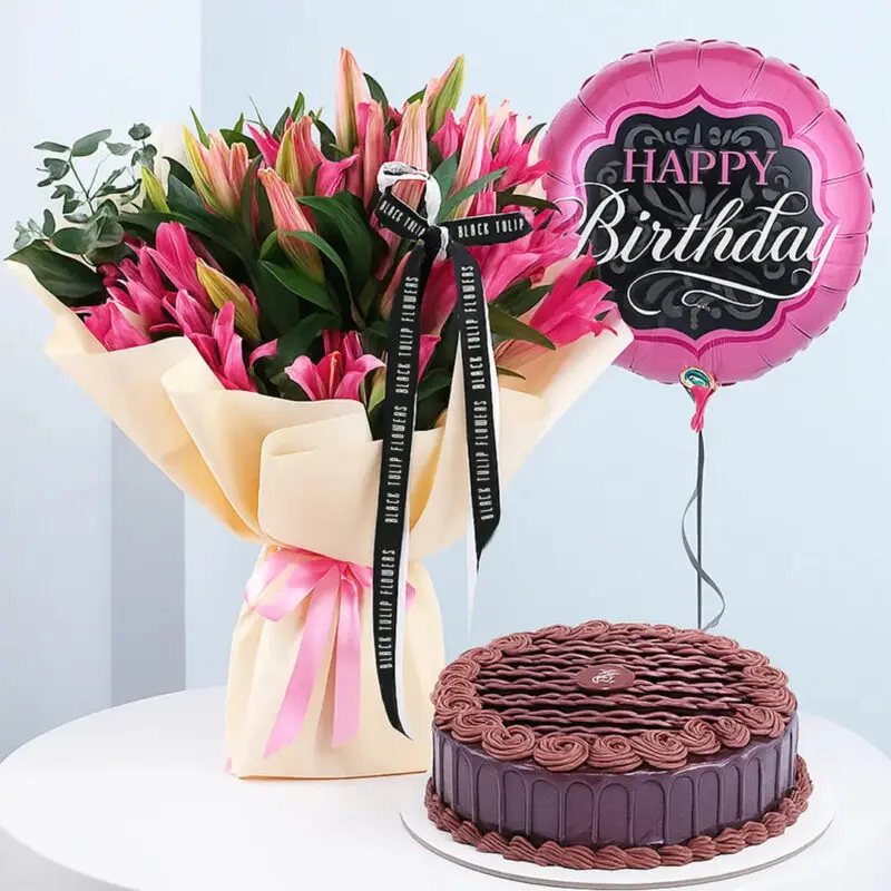 lilies-birthday-combo-10-25 A birthday combo featuring a bouquet of pink lily flowers wrapped in light yellow wrap with a pink ribbon, a round pink "Happy Birthday" balloon and a round chocolate cake with chocolate frosting.