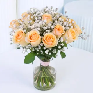 20 soft peach roses with white baby's breath arranged in a clear glass vase with a light pink ribbon.