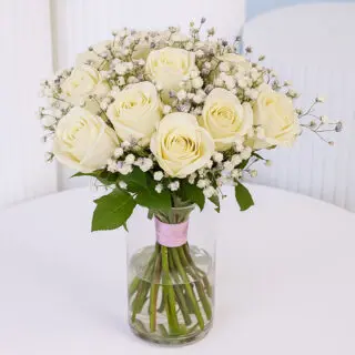 White rose floral arrangement in a glass vase with white baby’s breath tied with a pink ribbon.