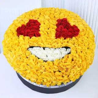 Emoji-inspired floral arrangement with yellow roses forming a round yellow face, red rose heart-shaped eyes, and a white rose smile, presented in a black round box.