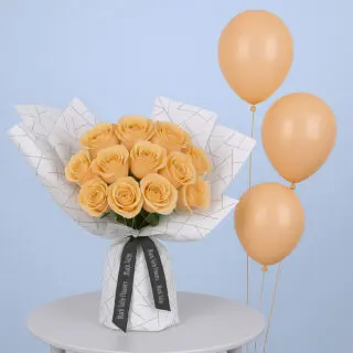 A bouquet of peach roses arranged in a white wrap with a black geometric design. It is tied with a black ribbon accompanied with peach-colored balloons.