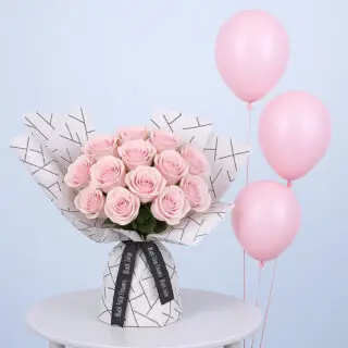 A bouquet of light pink roses wrapped in white paper with a black geometric pattern along with pink balloons.