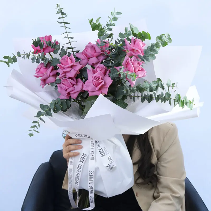 purple-roses-&-eucalyptus-bouquet_2026 A person sitting in a chair, holding a large bouquet of fresh lavender roses and green eucalyptus leaves, wrapped in white and tied with a white ribbon.