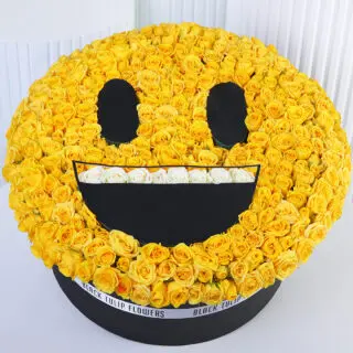 Floral arrangement shaped like a yellow grinning emoji face, made with yellow roses, black round eyes, a wide black smile, and white roses representing teeth.