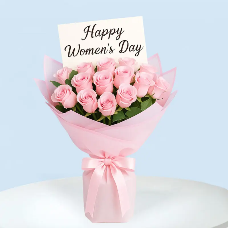 womens-day-rose-bouquet-01 A bouquet of soft pink roses wrapped in layers of light and dark pink paper and tied with a light pink satin ribbon, with a message card that reads "Happy Women's Day".