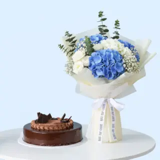 A bouquet featuring blue hydrangeas, white roses, baby's breath, and eucalyptus, wrapped in white and tied with white ribbon. A rich chocolate cake with a glossy glaze and chocolate frosting is adorned with chocolate bits.