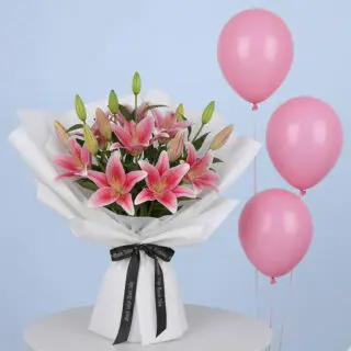 A vibrant pink lily bouquet with multiple blooming lilies and green buds, wrapped in white with a black branded ribbon and three pink balloons.