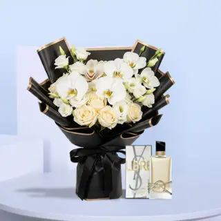 Black-wrapped bouquet of white phalaenopsis orchids, cream roses, and lisianthus with a satin ribbon, displayed beside a YSL Libre perfume box and bottle.