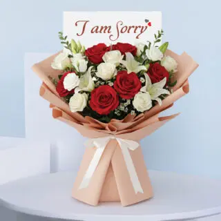 I am Sorry bouquet with red and white roses, white lilies, and tanacetum, wrapped in peach paper and tied with a white ribbon.