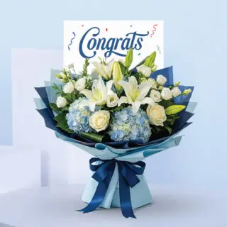 Blue hydrangeas, white lilies, and white lisianthus arranged with lush greens, styled in light-blue and navy layers, tied with a satin bow, and paired with a “Congrats” card.