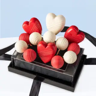 Red and white heart-shaped cakesicles and assorted chocolate truffles in a clear gift box with black ribbon.