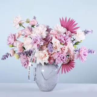 Light pink and purple roses with lisianthus and premium fillers in a marble-effect glass vase arrangement, finished with a white ribbon.
