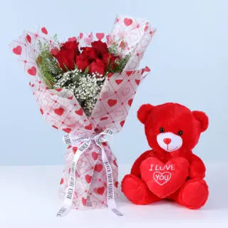 Bouquet featuring seven red roses styled with gypsophila and lepidium greenery in Valentine heart-print wrapping alongside a red teddy.