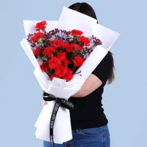 Bouquet of 20 red carnations and wax flower stems in white wrap, finished with a black ribbon.