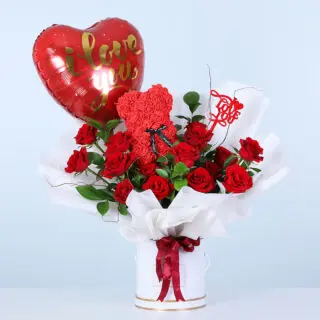 Red roses and green foliage in a white box, finished with a teddy bear and heart-shaped "I Love You" balloon.