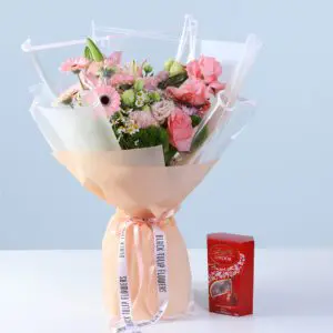 Bouquet of pink roses, gerberas, lilies, and seasonal blooms in soft peach wrapping, finished with a ribbon bow and paired with a Lindt Lindor chocolate box.