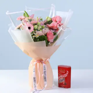 Bouquet of pink roses, gerberas, lilies, and seasonal blooms in soft peach wrapping, finished with a ribbon bow and paired with a Lindt Lindor chocolate box.