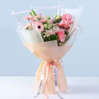 Bouquet of pink gerberas, roses, lilies, carnations, and seasonal fillers in peach and transparent wrapping, finished with a ribbon.