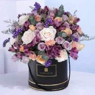 A purple-toned mix of fresh flowers elegantly arranged in a decorative black round floral box.