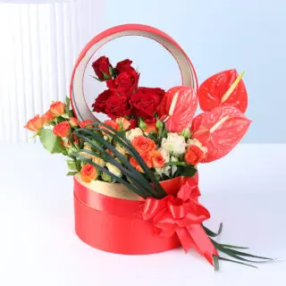 Red roses, orange spray roses, and red anthuriums in a round red flower box with gold detailing, finished with a ribbon bow.