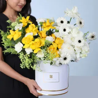 A white round box containing yellow roses, white roses, white carnations, white gerbera daisies, baby white orchids, solidaster flowers, and gypsophila arranged with decorative foliage in a bright and elegant floral arrangement.