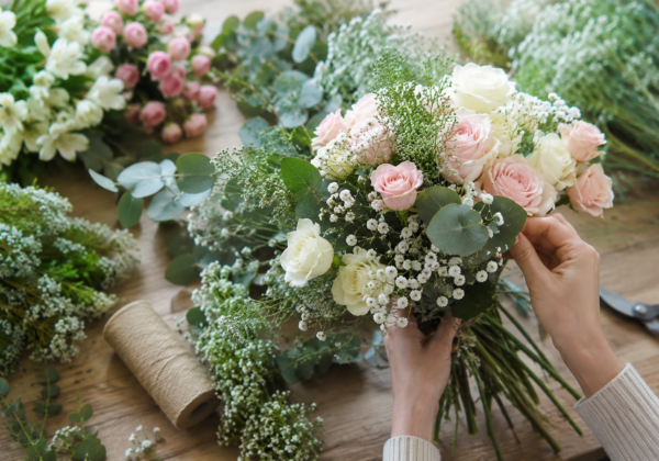 Hands arranging a bouquet of pink and white roses, baby's breath, and greenery on a wooden table with floral supplies and tools nearby.